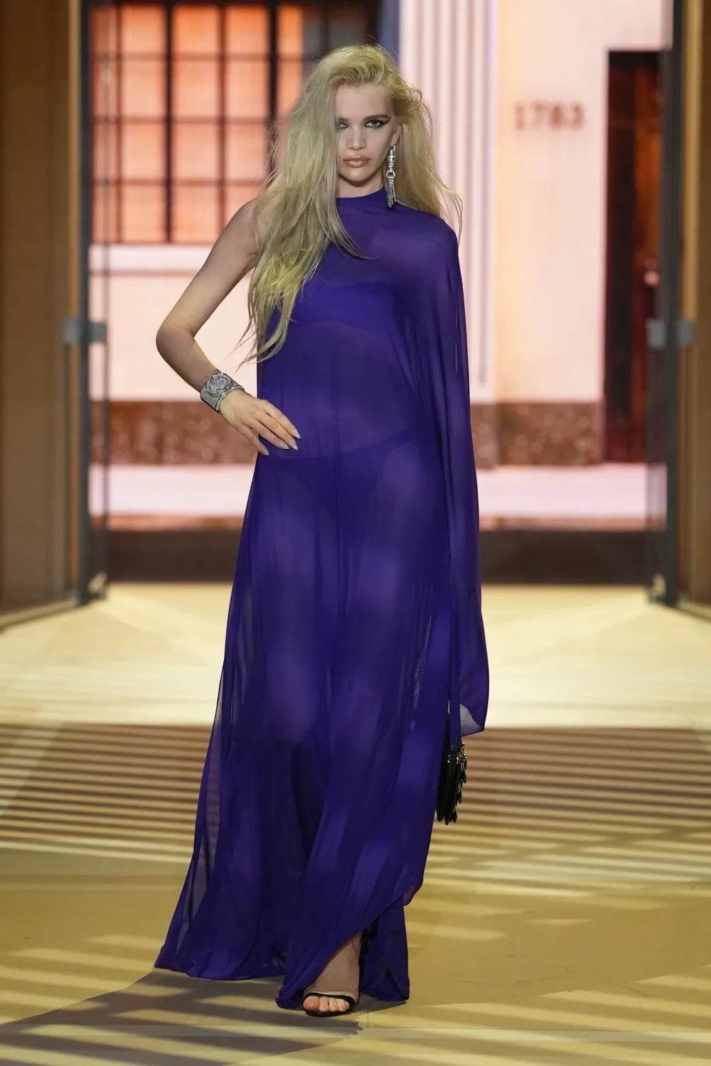 Modelo luciendo un vestido largo y fluido en tono morado intenso, de corte asimétrico con una sola manga y transparencias sutiles, fotografiada por Alessandro Fucioni.
