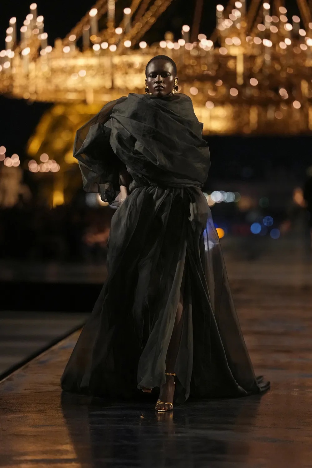 Primer plano de una modelo desfilando con un vestido negro estructurado con grandes volúmenes drapeados en la parte superior y pendientes dorados llamativos.