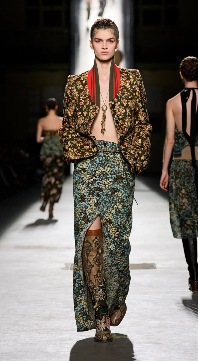 Modelo con un conjunto de chaqueta corta y falda larga a juego con estampado floral azul y verde, luciendo botas altas de piel con estampado de serpiente y aberturas frontales.