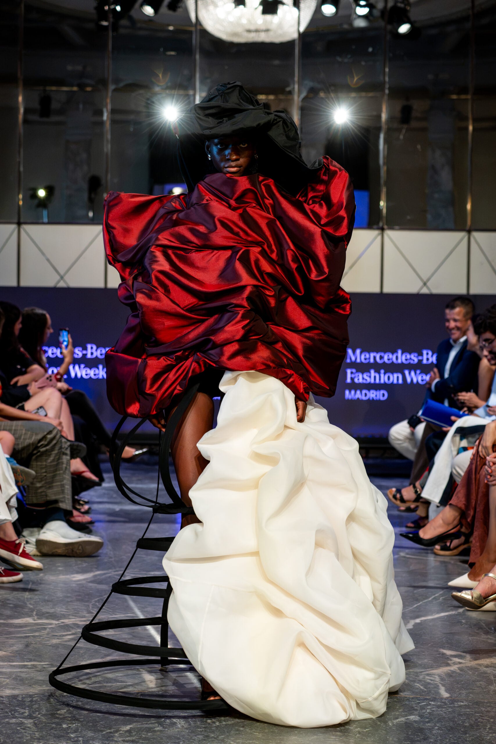 Modelo desfilando con un diseño vanguardista y dramático compuesto por un corpiño voluminoso de tafetán rojo brillante, tocado negro texturizado y una estructura de miriñaque inferior que sostiene una falda blanca drapeada.