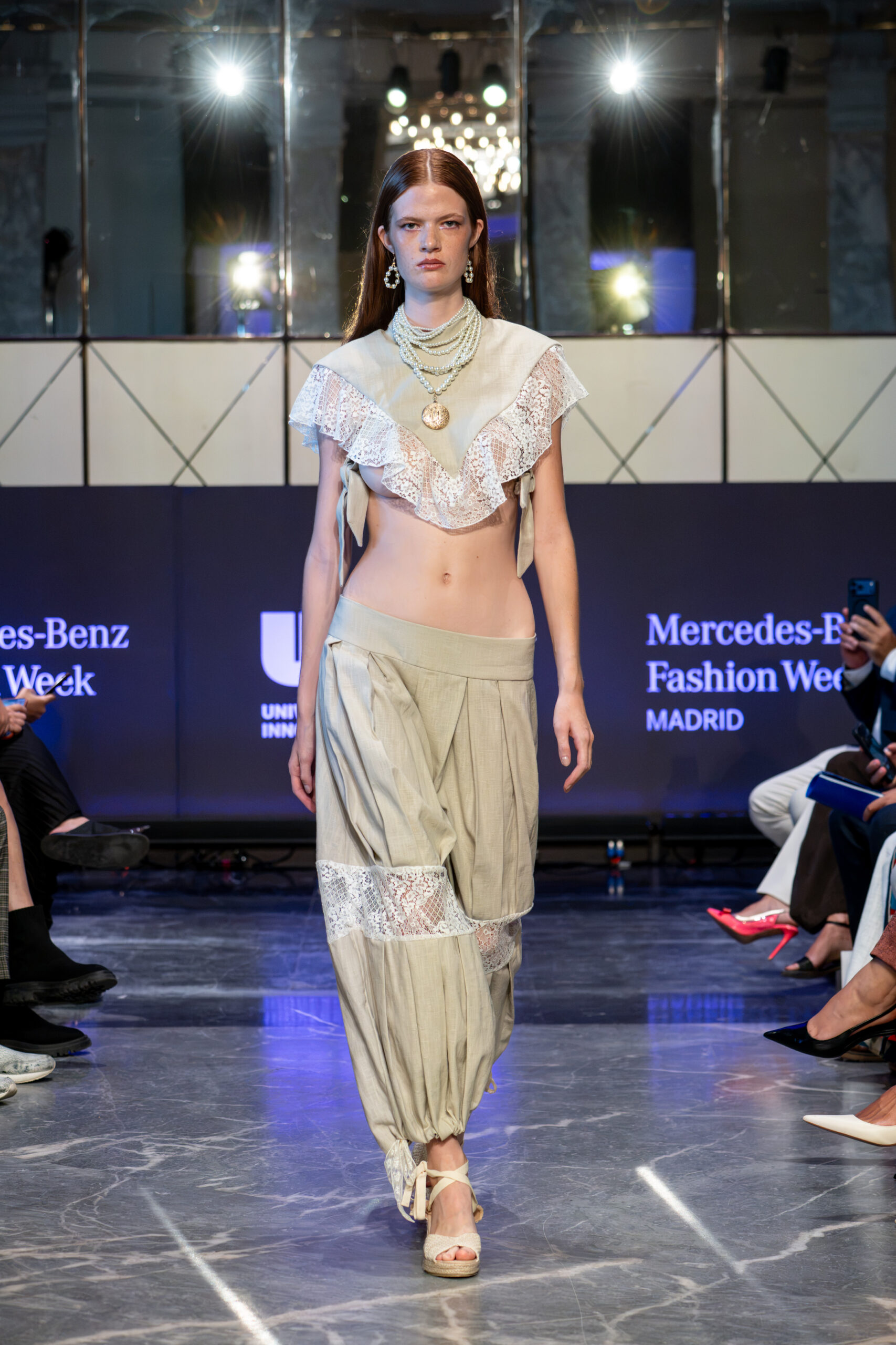 Modelo con cabello pelirrojo desfilando con un conjunto en tono gris claro compuesto por un top corto con volantes de encaje, collares de perlas superpuestos y pantalones bombachos anchos con inserciones de encaje.