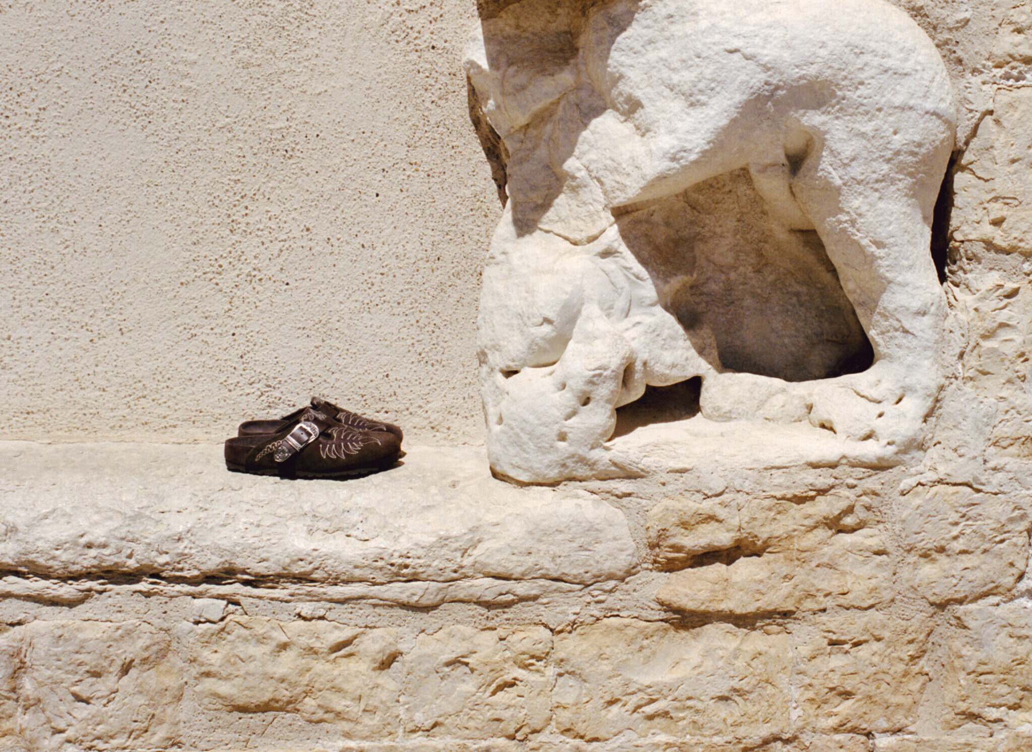 Primer plano de un par de zuecos Boston de ante marrón oscuro con bordados florales detallados, colocados sobre la cornisa de un muro de piedra antigua.