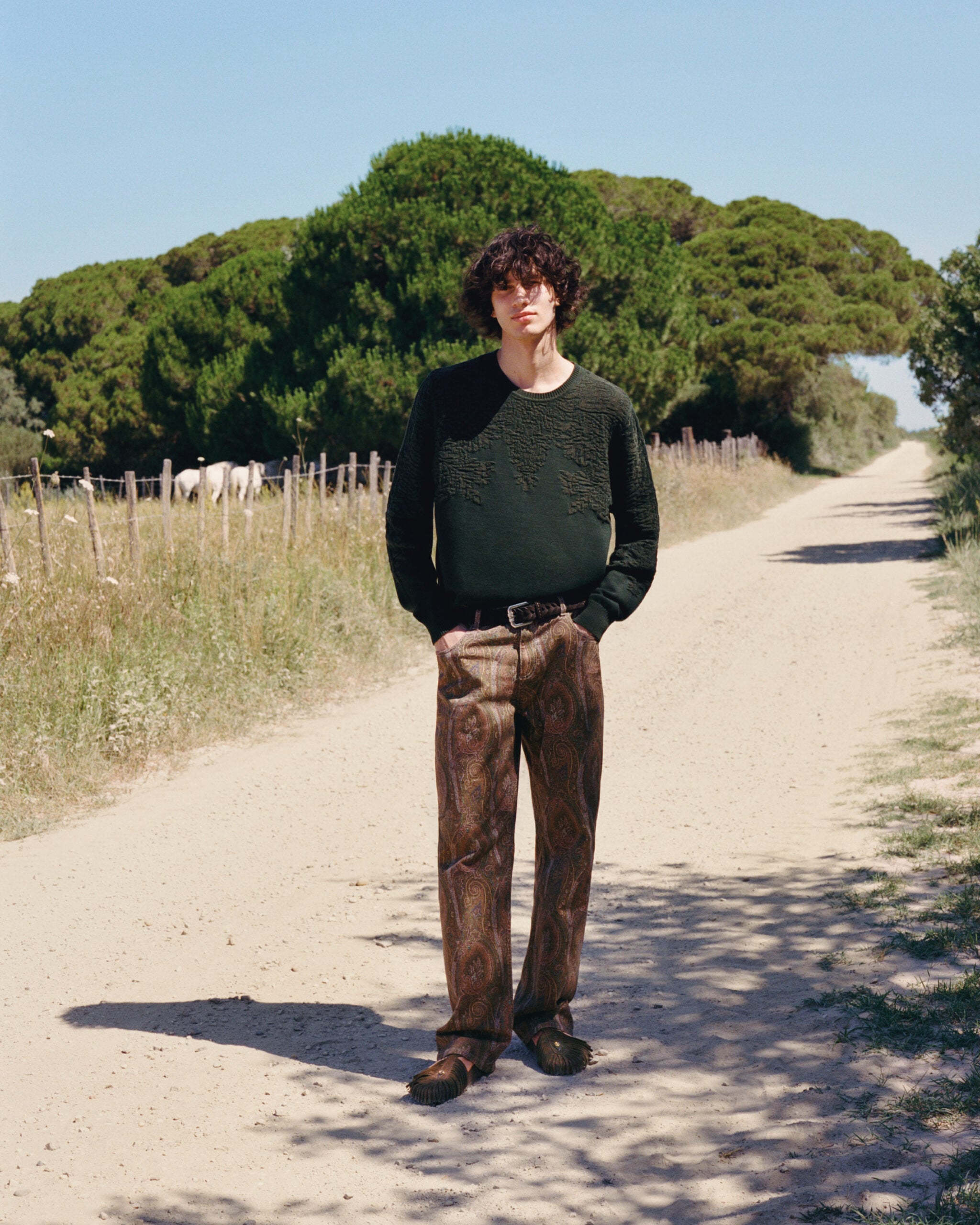 Modelo masculino parado en un camino rural, vistiendo un suéter de punto verde oscuro con texturas de bordado, pantalones rectos con estampado Paisley y sandalias de ante con flecos.