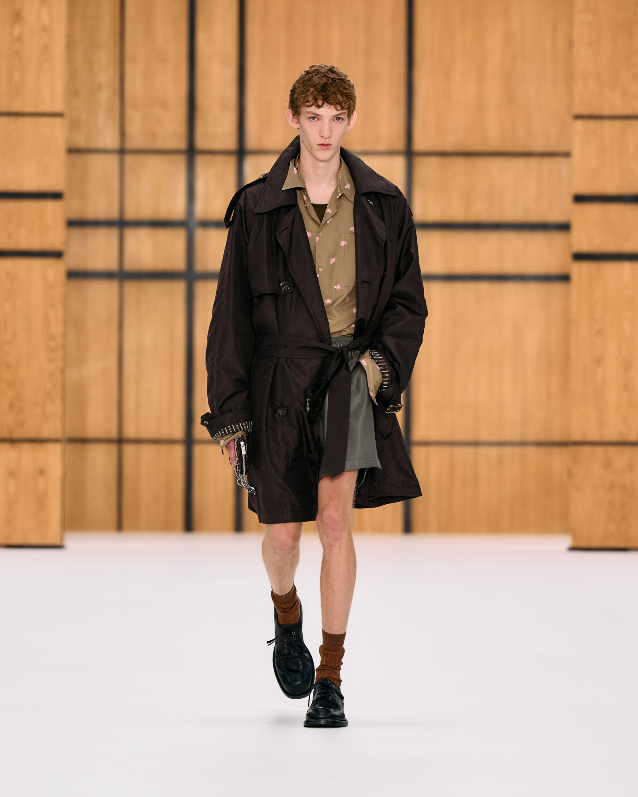 Modelo masculino con un trench coat negro ligero, camisa estampada caqui, bermudas oscuras y botas de combate con calcetines marrones.