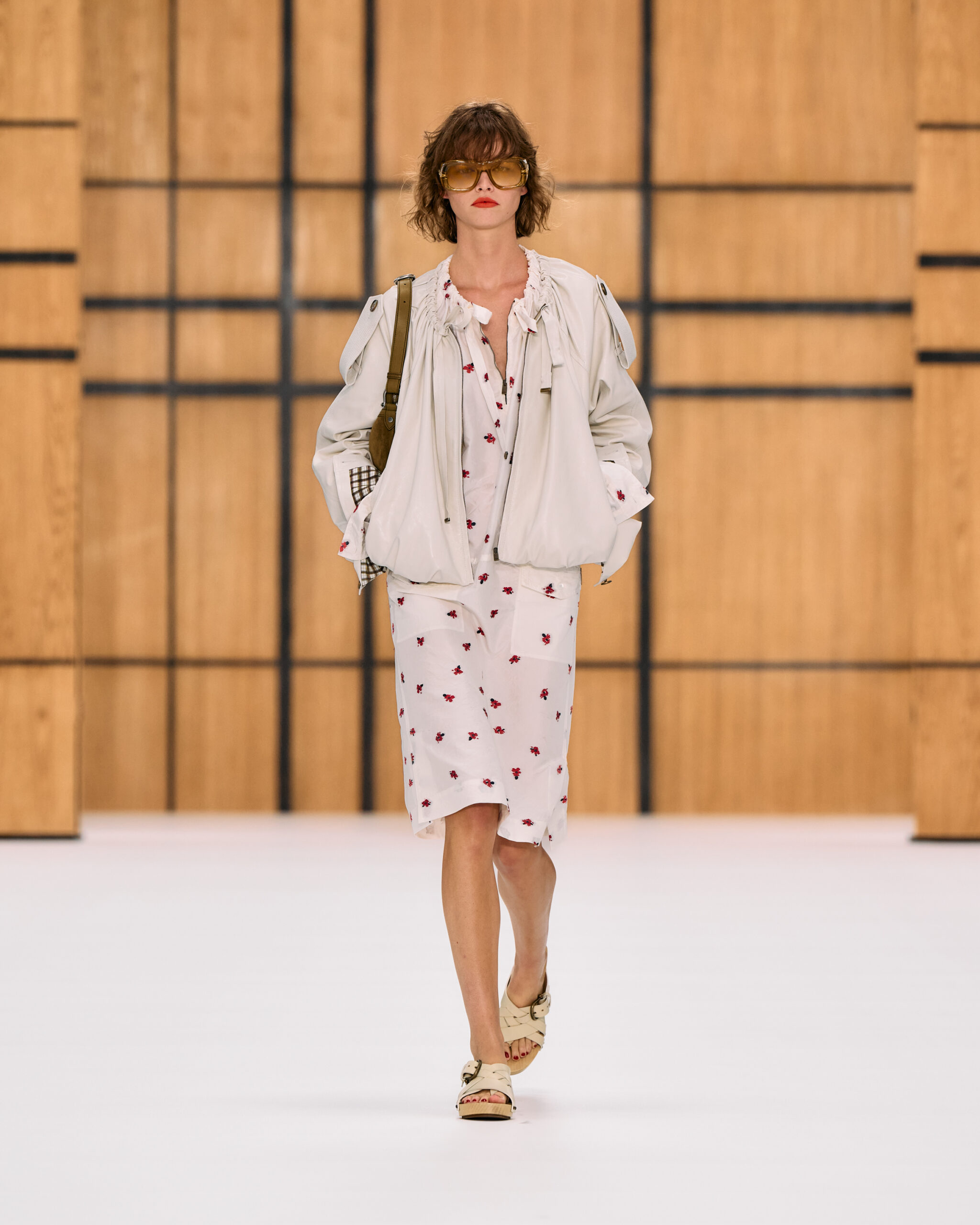 Modelo con un vestido midi blanco con estampado floral rojo, chaqueta corta beige estructurada, lentes de sol y sandalias planas de tiras.