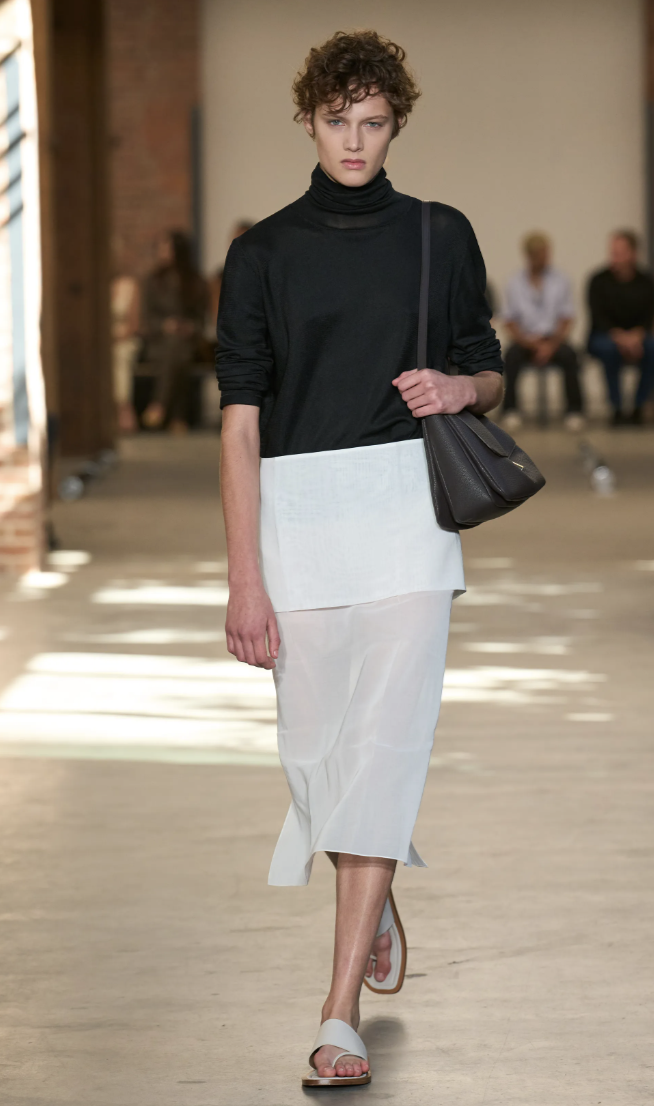 Modelo con un suéter negro de cuello alto, falda blanca midi semitransparente, bolso de mano oscuro y sandalias planas.