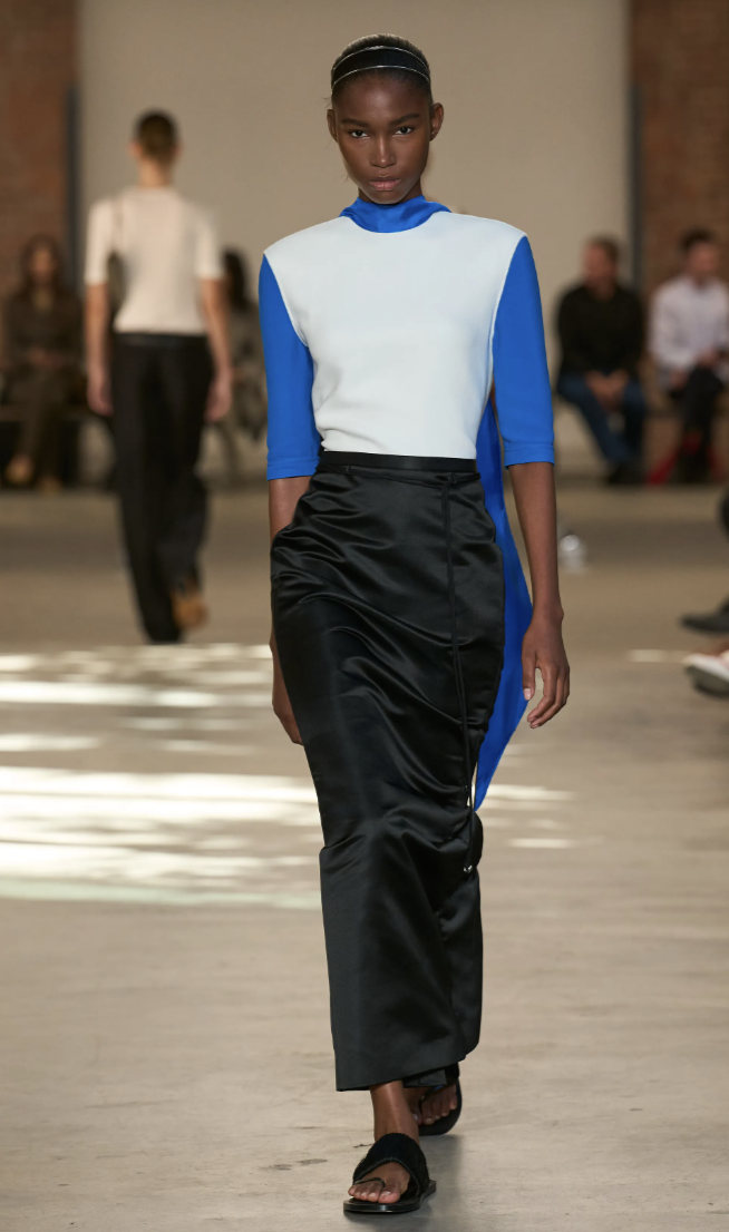 Modelo con blazer negro estructurado sobre top blanco, falda midi negra con un fular o detalle en azul vibrante y bolso de mano.
