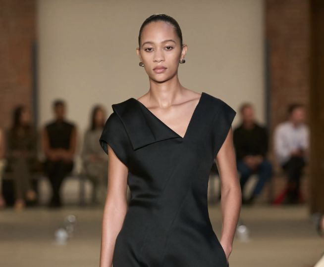 Modelo con un vestido largo negro de diseño minimalista con cuello asimétrico estructurado y sandalias finas de tiras.