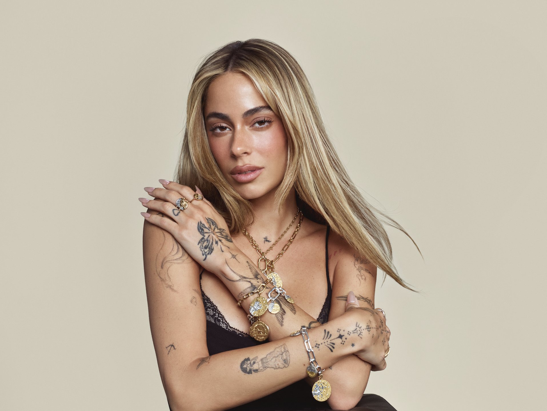 Tini con el cabello rubio suelto con mechas más claras, vistiendo un top negro de tirantes y luciendo múltiples collares, pulseras y anillos de la colección Pandora Talisman con dijes del zodiaco.