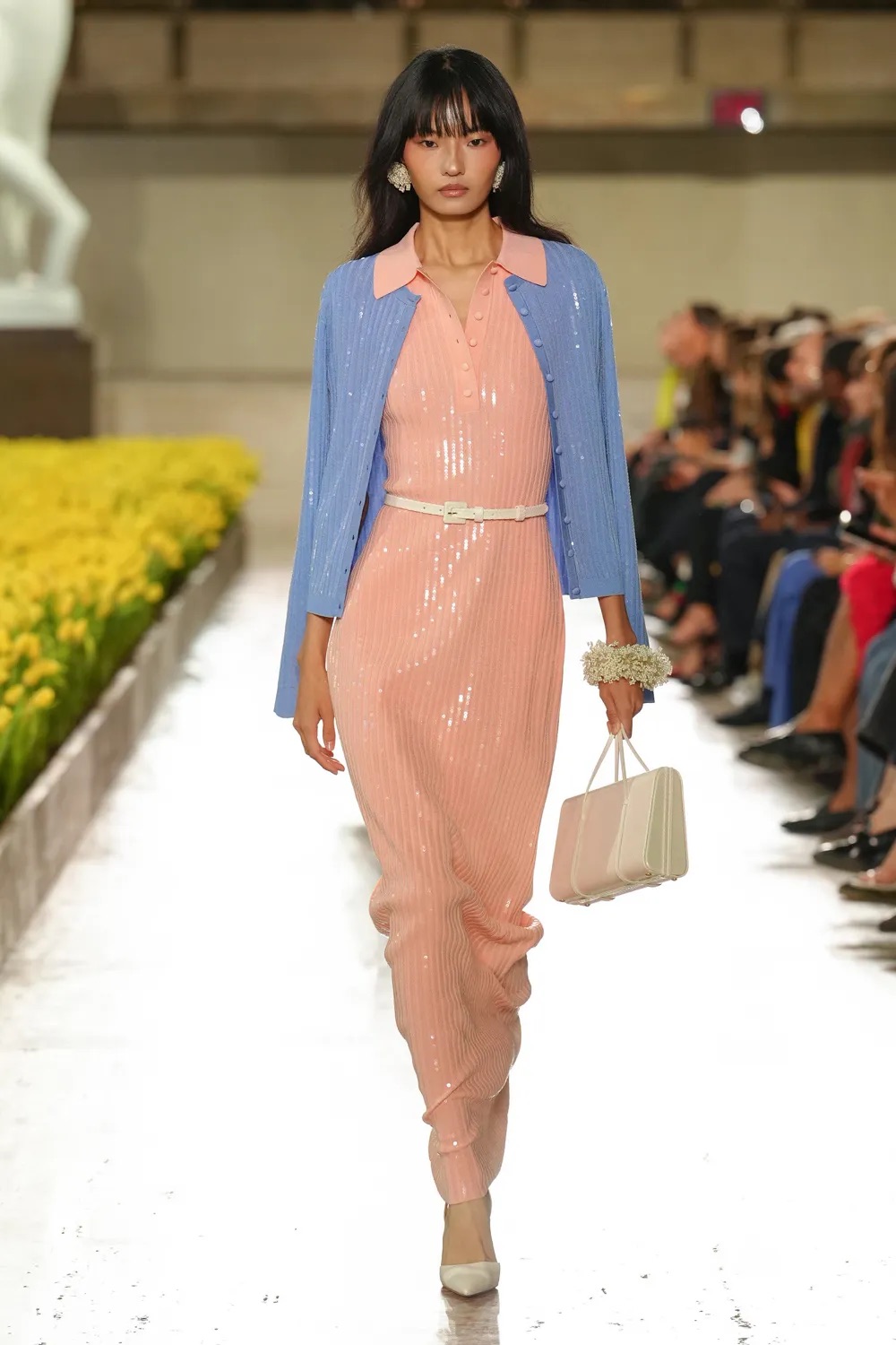 Modelo desfilando con un vestido largo de punto acanalado en tono rosa pastel con cuello tipo polo, bajo una chaqueta de punto azul claro con detalles brillantes, un cinturón blanco fino y bolso de mano a juego.