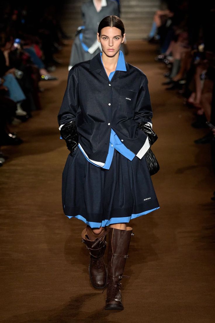 Modelo en pasarela con un conjunto superpuesto de camisa azul, chaqueta oscura estructurada, falda amplia y botas biker de piel marrón con múltiples correas.