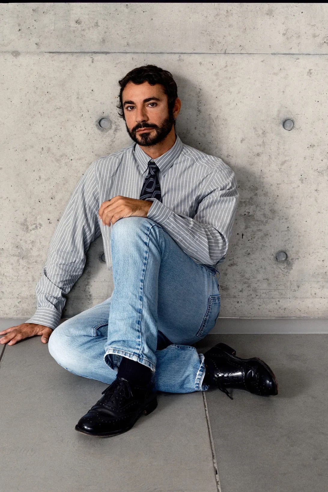 Retrato de Dario Vitale sentado en el suelo apoyado contra una pared de concreto, vistiendo una camisa de rayas, corbata, jeans claros y zapatos negros de charol.