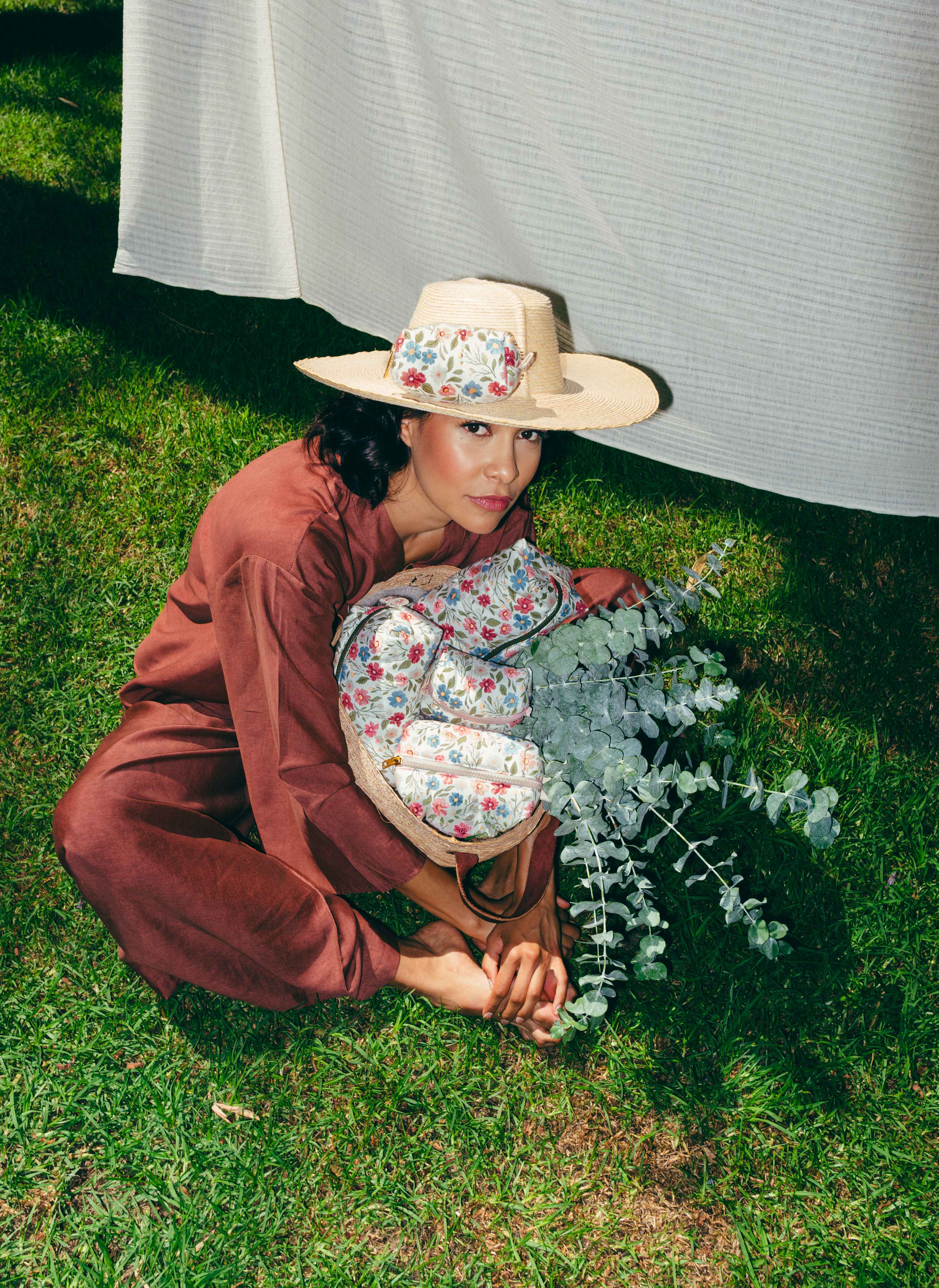 Modelo con sombrero de paja y conjunto en tono terracota, sentada en el césped mientras sostiene una cesta con varios neceseres de estampado floral y ramas de eucalipto.