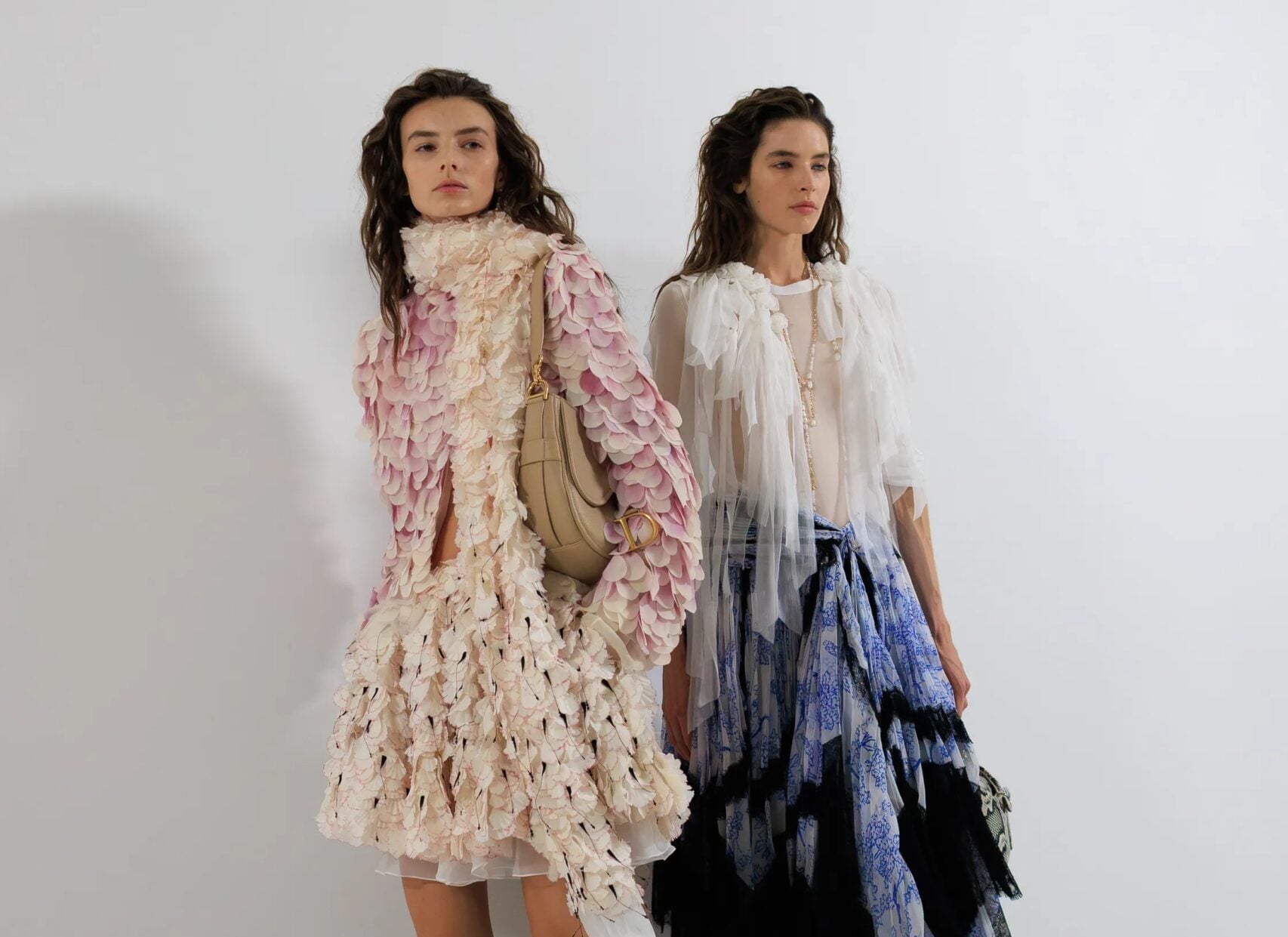 Dos modelos posando en el backstage del desfile, una luciendo un vestido corto cubierto de aplicaciones texturizadas en tonos degradados de rosa y blanco, y la otra con una blusa blanca vaporosa y una falda larga estampada en tonos azules con detalles de encaje negro.