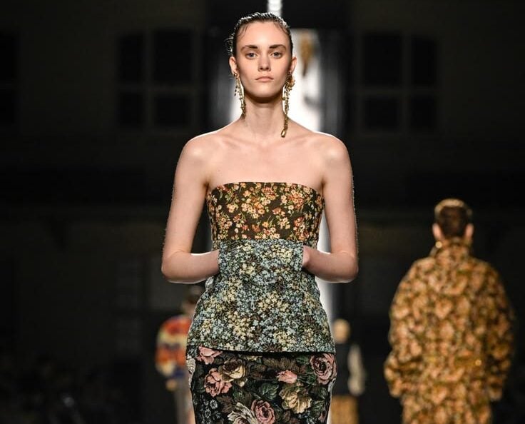 Modelo en la pasarela portando un top palabra de honor con estampado floral multicolor y una falda larga recta de terciopelo con grandes rosas estampadas.