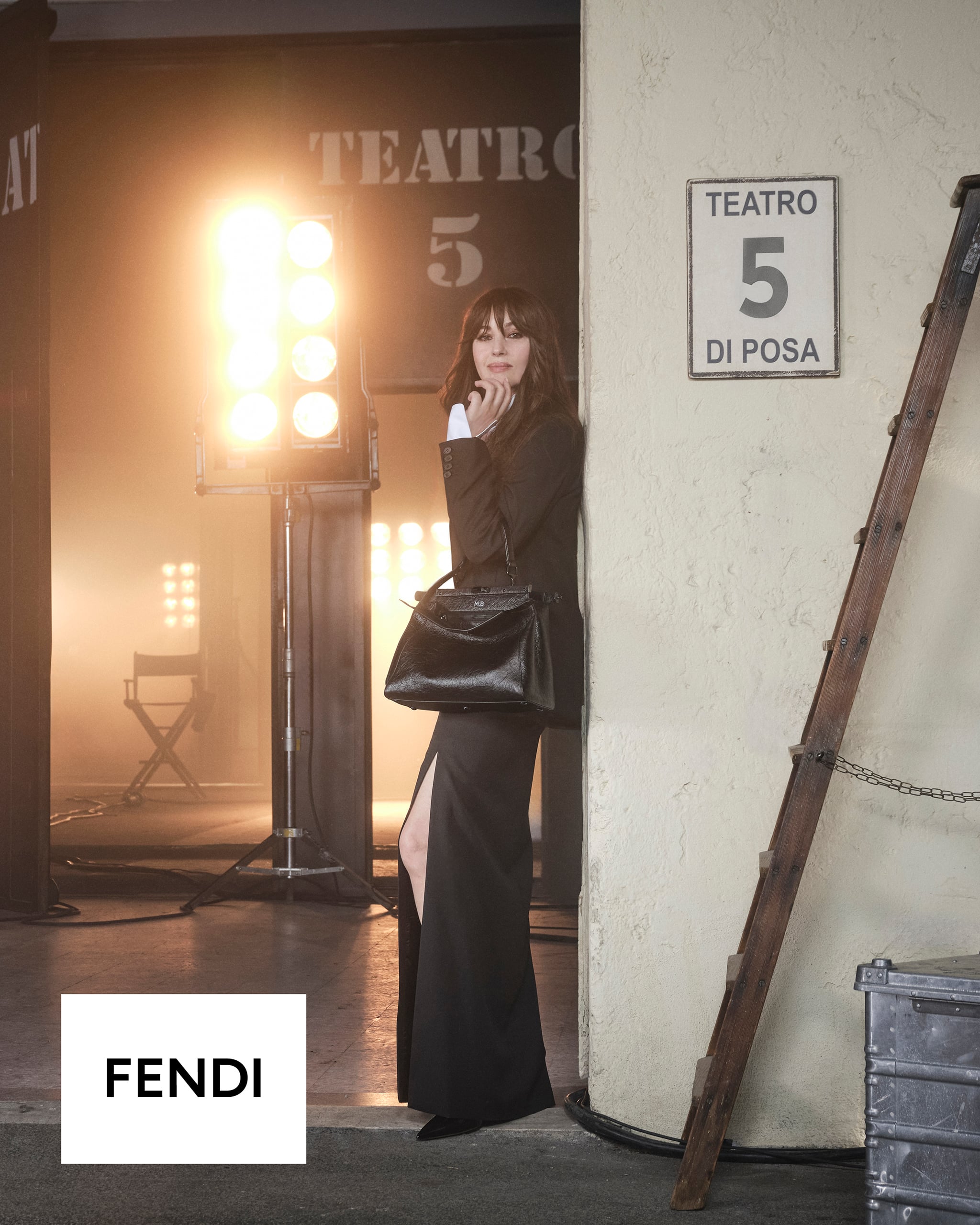 Monica Bellucci posa en un estudio con iluminación cinematográfica, luciendo un elegante conjunto negro con abertura lateral y sosteniendo el bolso Peekaboo de FENDI.