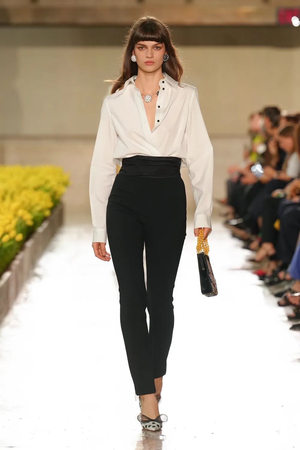 Modelo con una camisa blanca formal de cuello abierto con solapa cruzada, pantalones negros ajustados de talle alto con faja de satén y zapatos de tacón transparente con detalle oscuro.