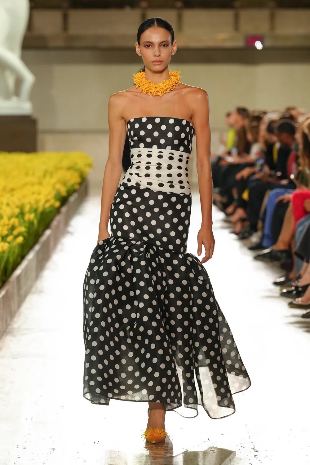 Modelo luciendo un vestido strapless de tubo con estampado de lunares (polka dots) en blanco y negro, un fajín de lunares invertidos en la cintura y una falda con volantes translúcidos en el bajo.