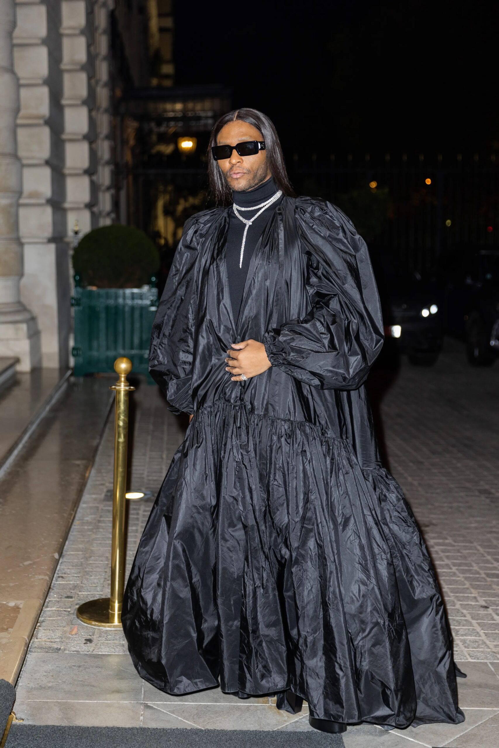 Law Roach camina por una calle nocturna luciendo un voluminoso abrigo o vestido largo de color negro con mangas abullonadas, gafas de sol oscuras y collares de diamantes en capas.