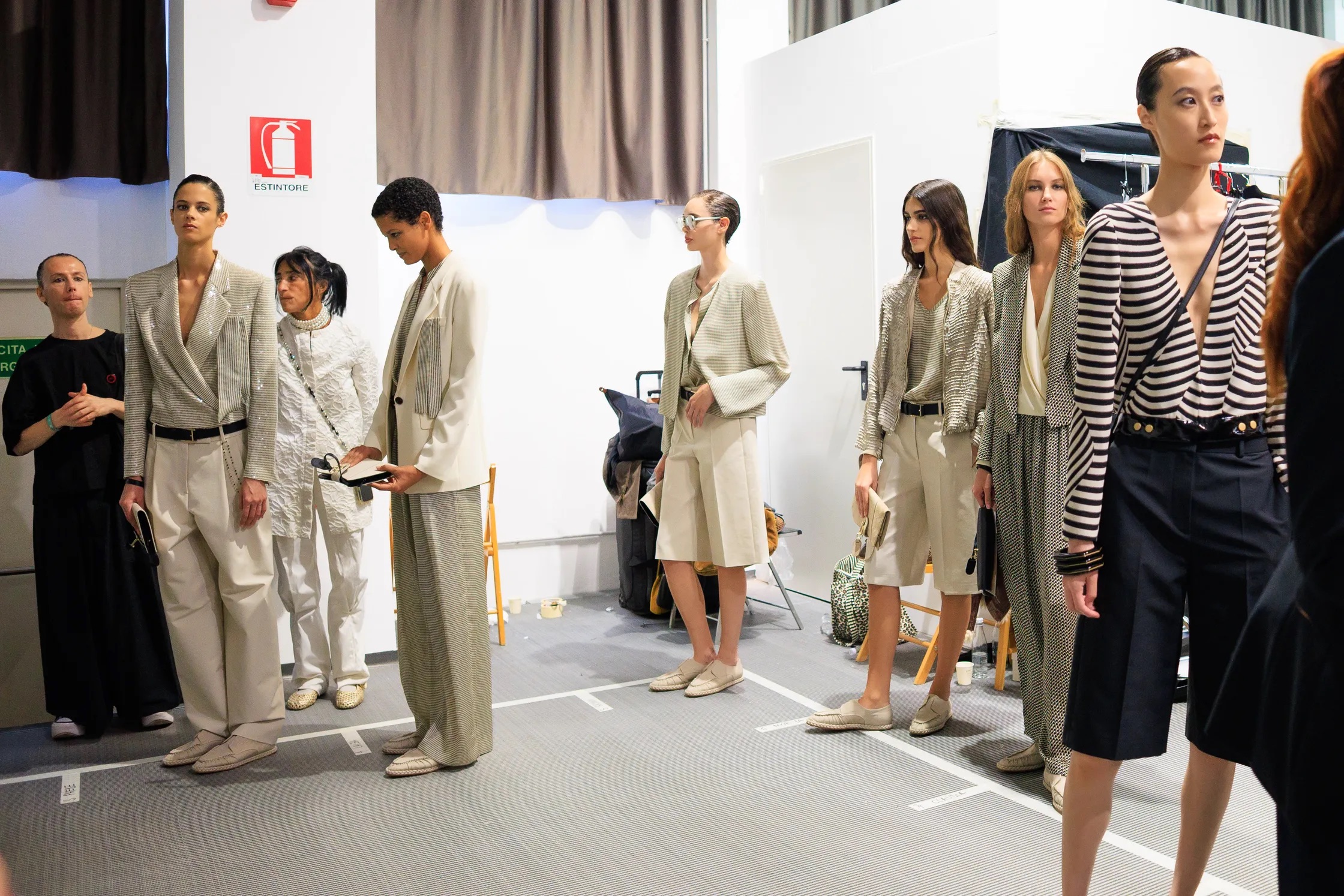 Modelos en el backstage preparándose con conjuntos fluidos en tonos neutros, chaquetas ligeras de corte sastre y pantalones anchos con cinturones finos, capturadas por Acielle StyleDuMonde