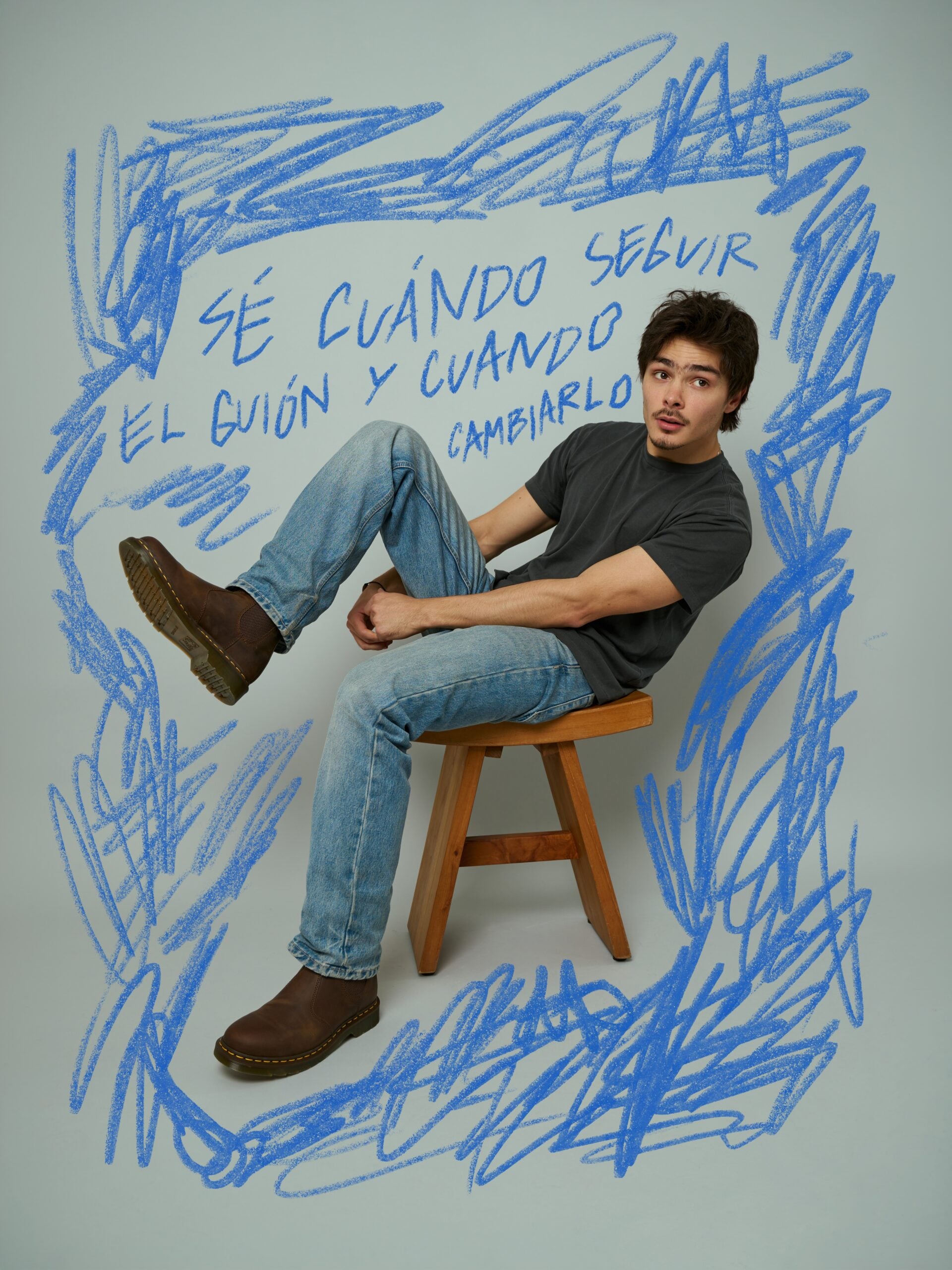 Joven con playera gris y jeans de mezclilla sentado en un banco de madera sobre un fondo decorado con un marco azul dibujado a mano y el texto "Sé cuándo seguir el guión y cuándo cambiarlo", vistiendo botines chelsea marrones.