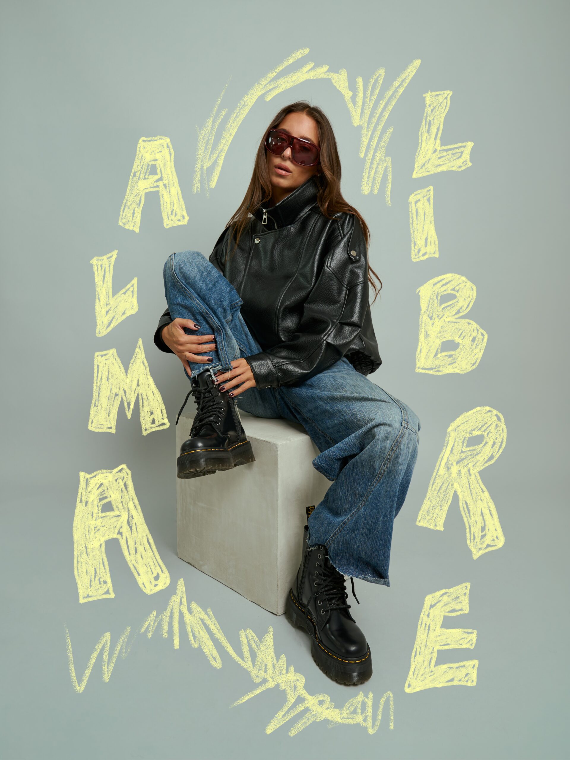 Modelo con chaqueta de piel negra, gafas de sol y jeans anchos sentada sobre un cubo, portando botas altas con plataforma y rodeada por letras amarillas dibujadas que forman la frase "El alma libre".