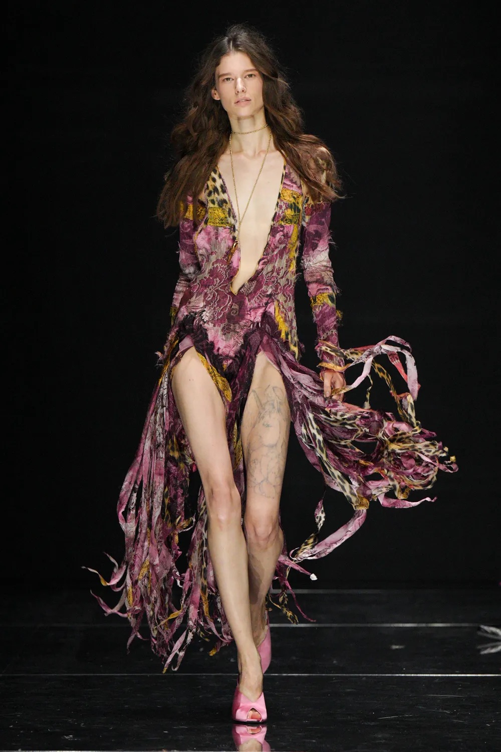 Modelo luciendo un vestido largo y fluido de escote pronunciado en V, con un diseño texturizado en tonos morados, burdeos y destellos dorados, adornado con tiras colgantes y flecos flotantes.