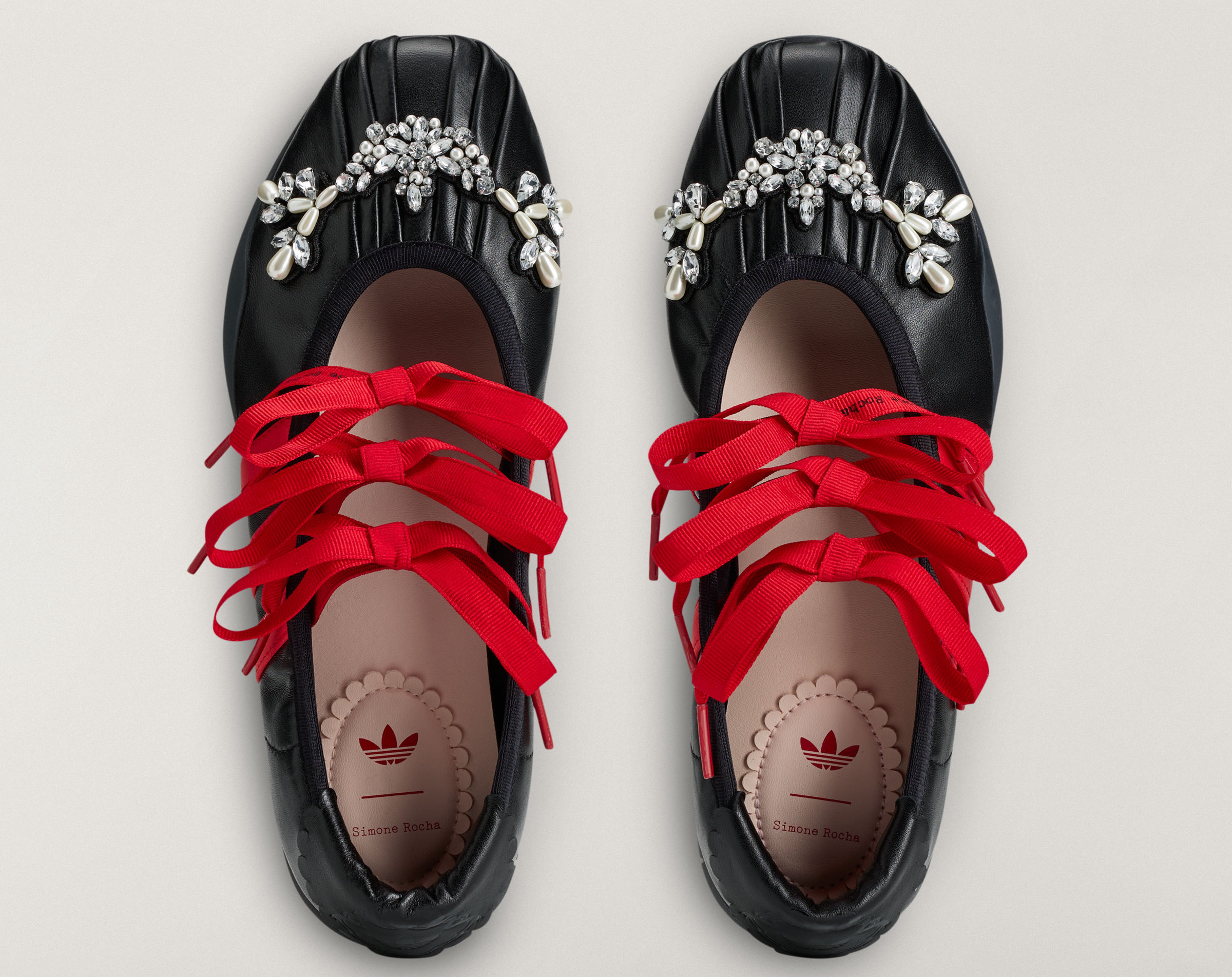 Vista superior de un par de zapatillas estilo bailarinas negras de piel plisada con grandes lazos rojos y un adorno floral de cristales y perlas en la puntera