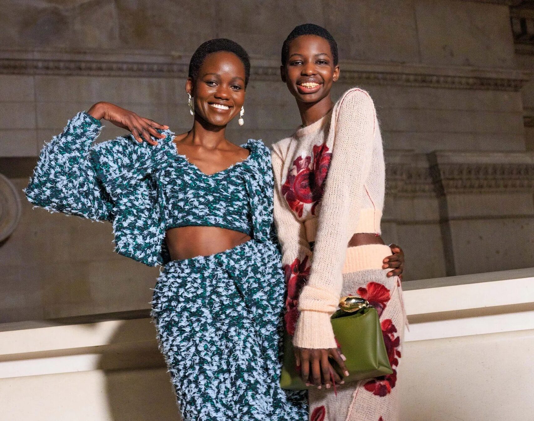 Dos modelos posando sonrientes en el backstage; la primera lleva un conjunto de dos piezas texturizado en tonos verde oscuro y azul, y la segunda un vestido de punto claro con detalles estampados y un bolso verde oliva.