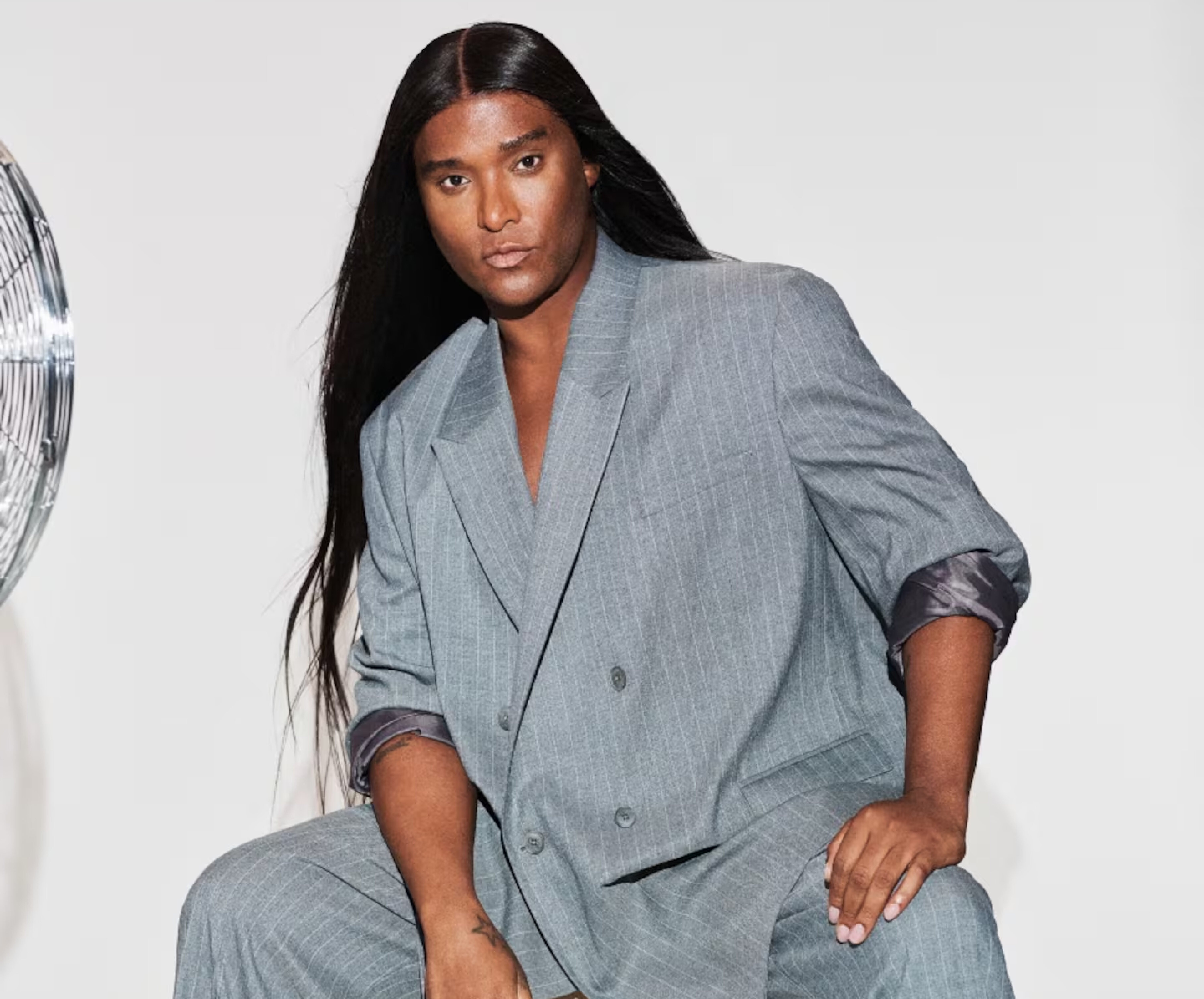Law Roach sentado sobre una caja de madera con el logotipo de ASOS, vistiendo un traje sastre gris de raya de tiza estilo oversize, botines negros y el cabello largo y lacio.
