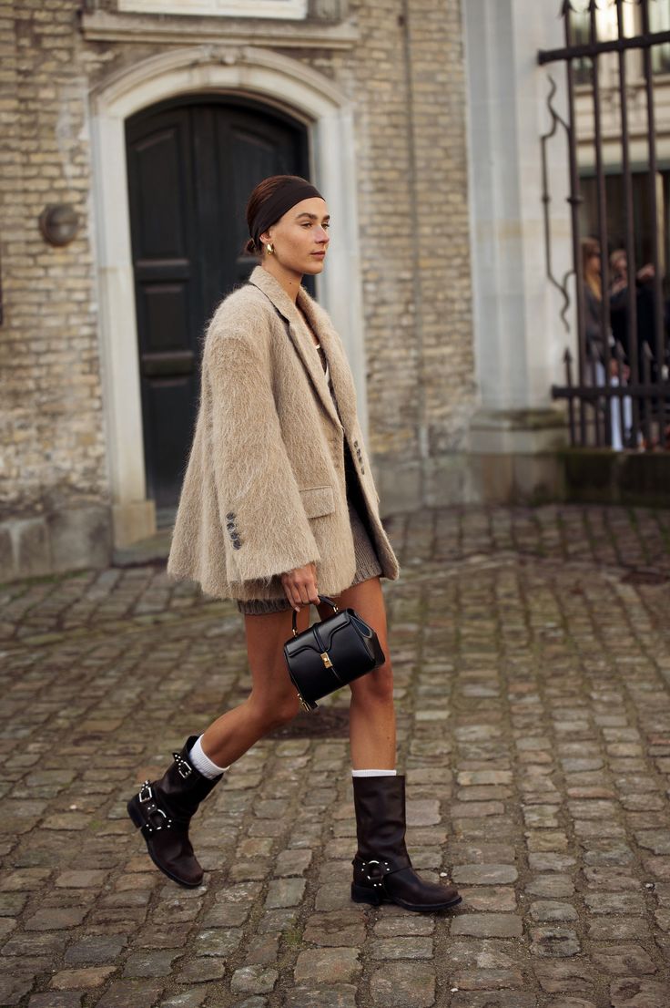 Mujer cruzando una calle empedrada luciendo un abrigo extragrande de efecto afelpado en tono beige, minivestido, calcetines blancos visibles y botas biker oscuras.