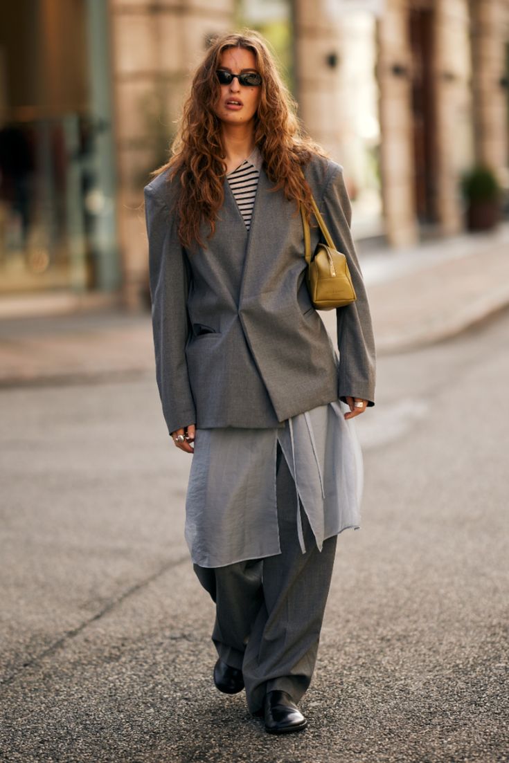 Mujer con melena ondulada luciendo un abrigo gris oversize, una falda de gasa gris translúcida sobre pantalones anchos a juego y un bolso de mano verde oliva.