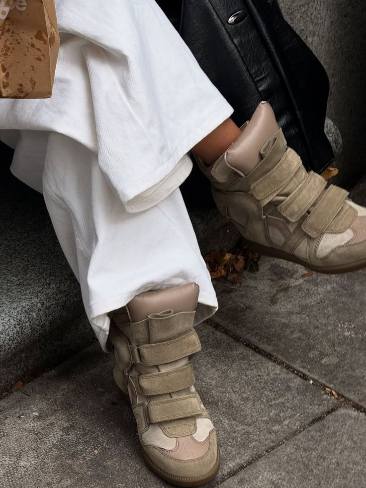 Primer plano de unos tenis de caña alta con cuña en tonos beige y marrón, provistos de múltiples tiras de velcro, combinados con pantalones blancos amplios.
