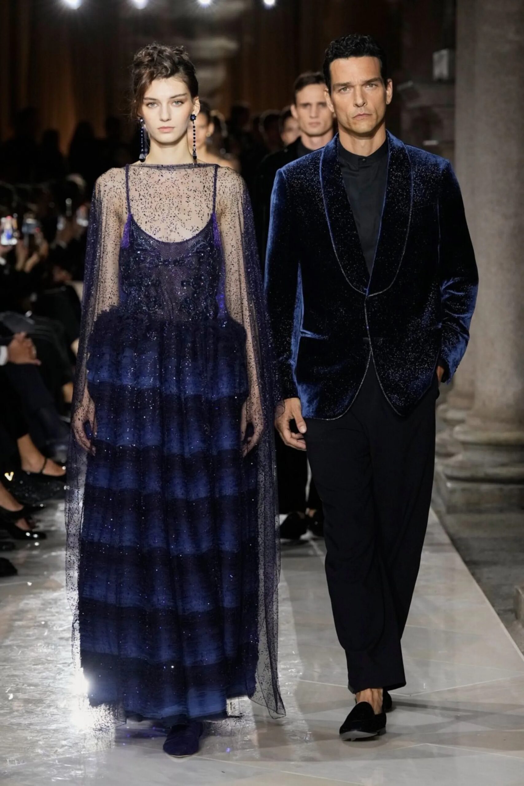 Modelos desfilando en la pasarela; una mujer con un vestido largo de tul azul con capa transparente y un hombre con traje de terciopelo azul marino.