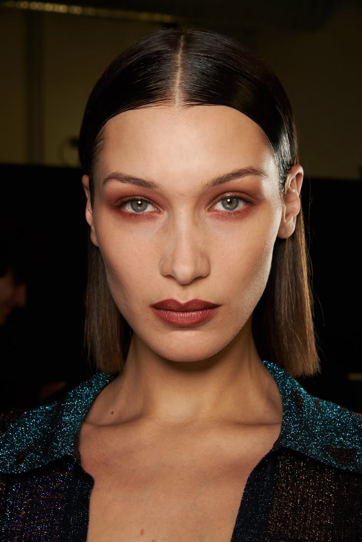 Primer plano de Bella Hadid con cabello lacio de raya al medio y efecto mojado, luciendo sombras en tonos terracota cálidos alrededor de los ojos y labios perfilados en un tono vino profundo.
