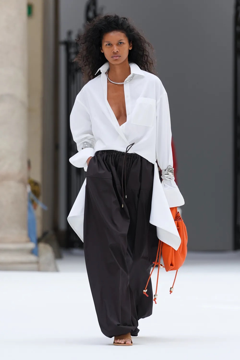 Modelo con una blusa blanca de cuello camisero amplio y escote pronunciado, combinada con pantalones oscuros tipo paracaídas con jareta y un bolso de mano en tono naranja vibrante.