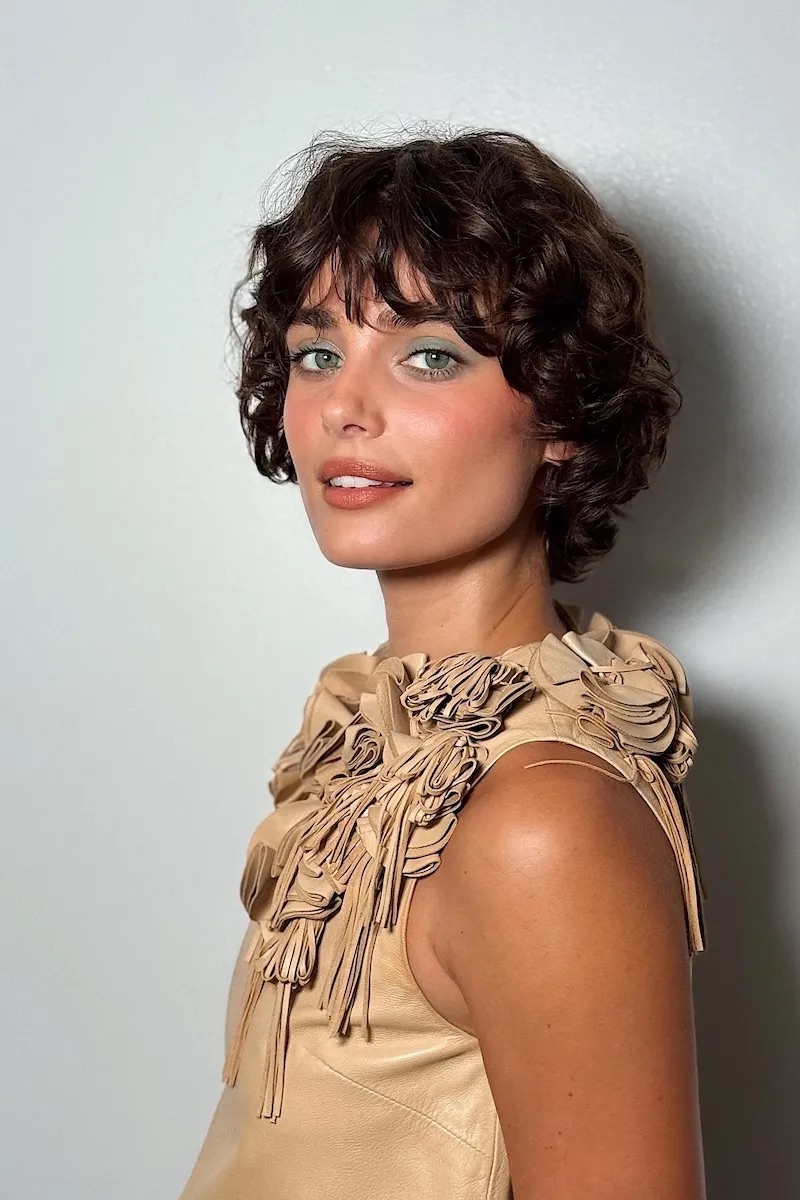 Taylor Hill con cabello corto rizado y flequillo, vistiendo una prenda estructurada con detalles florales en relieve, y luciendo sombras en un tono verde suave en los párpados.
