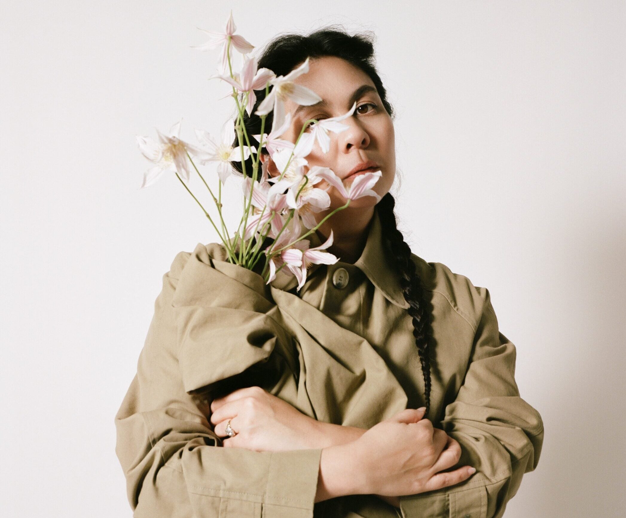 Retrato de Simone Rocha con un abrigo gabardina caqui, sosteniendo un ramo de flores delicadas cerca de su rostro y con el cabello trenzado.