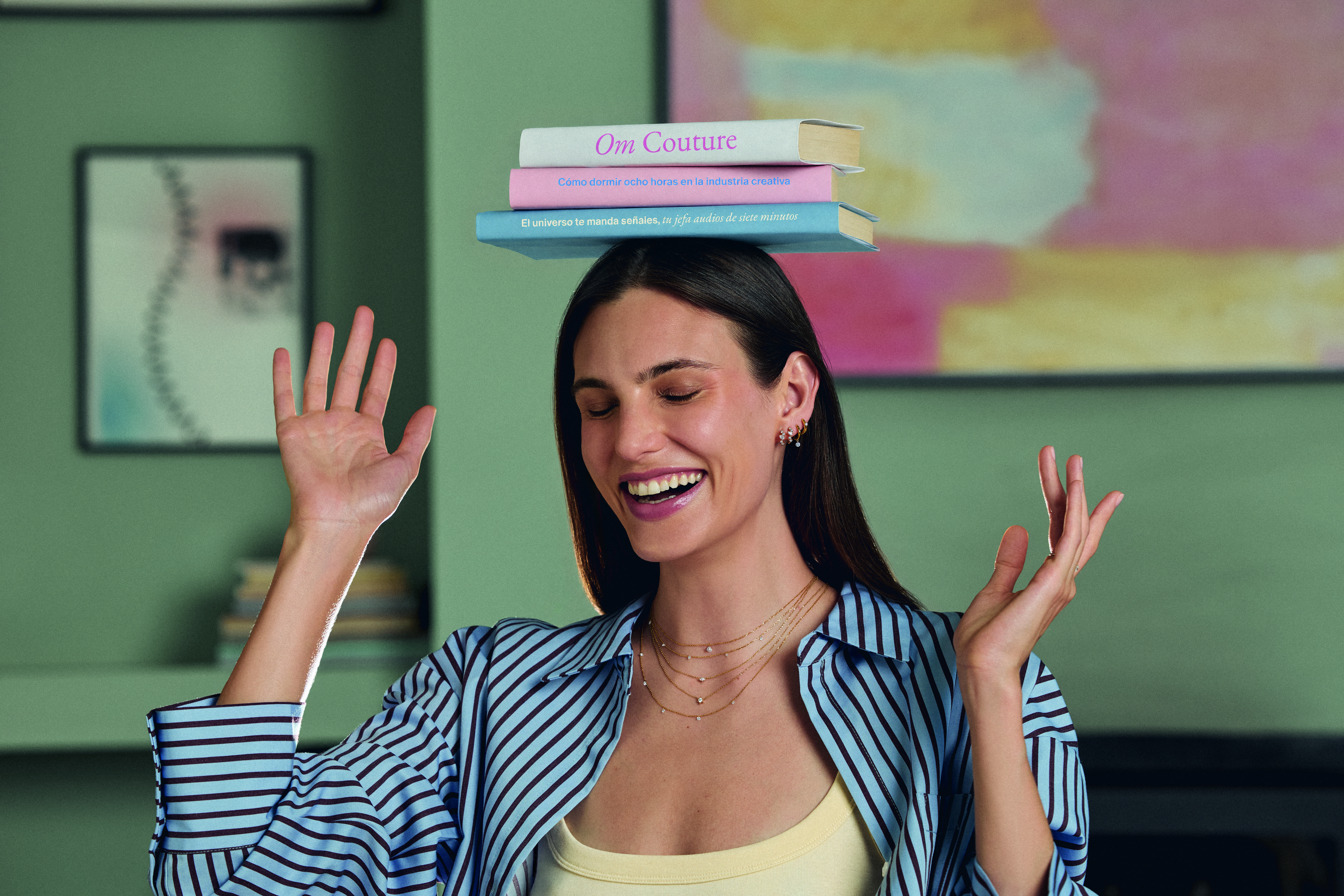 Modelo sonriendo con las manos levantadas y tres libros apilados equilibrados sobre su cabeza, luciendo una camisa de rayas azul y blanca y varias cadenas delicadas con diamantes en el cuello.