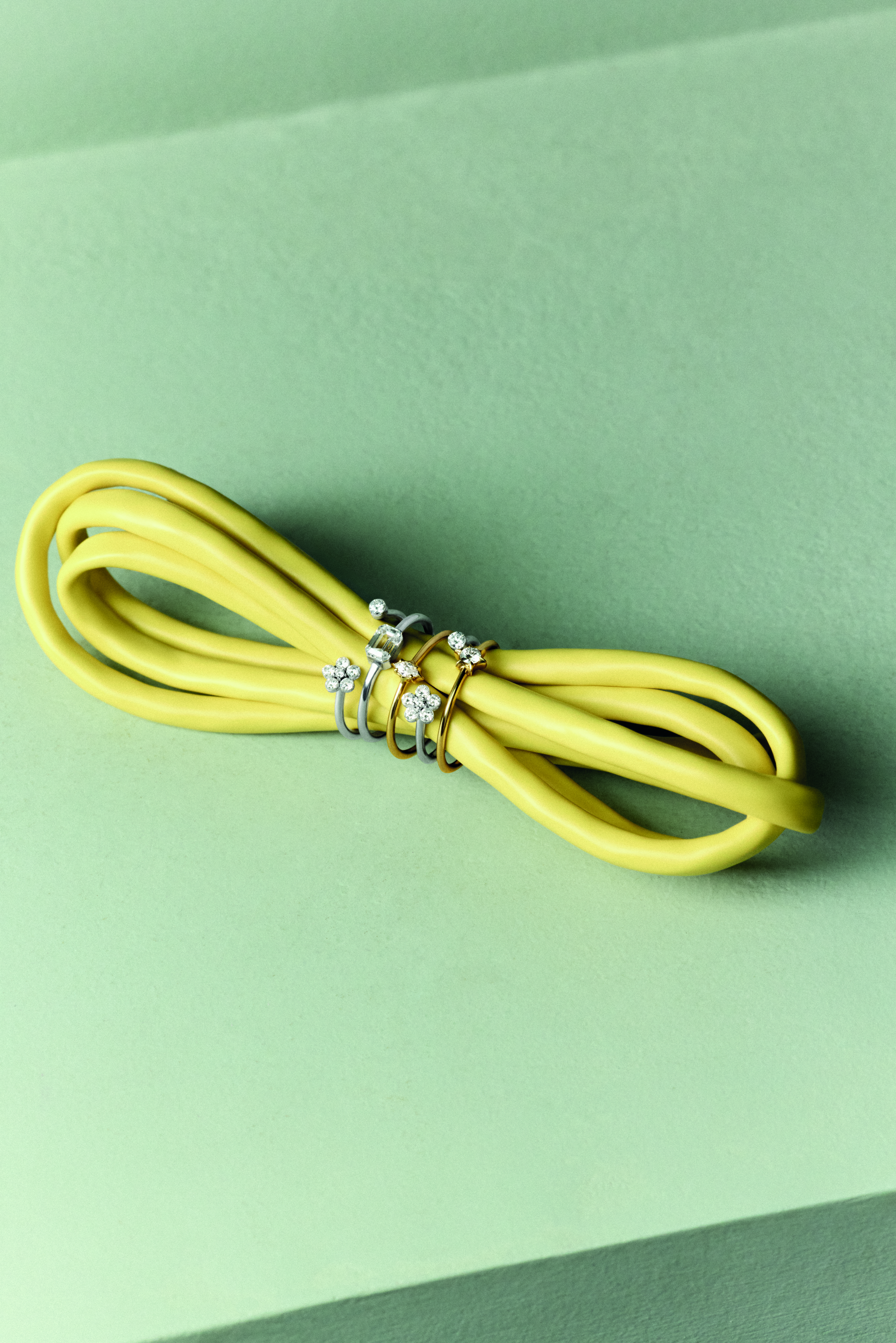 Cuatro anillos de oro y plata con engastes de diamantes en formas florales y geométricas, entrelazados alrededor de un cordón amarillo brillante sobre una superficie verde claro.