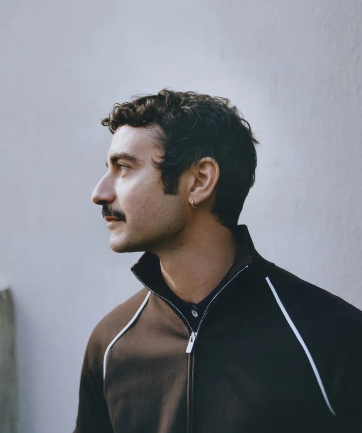Primer plano de perfil de Dario Vitale con cabello rizado corto, bigote y una chaqueta deportiva oscura con detalles blancos en los hombros.