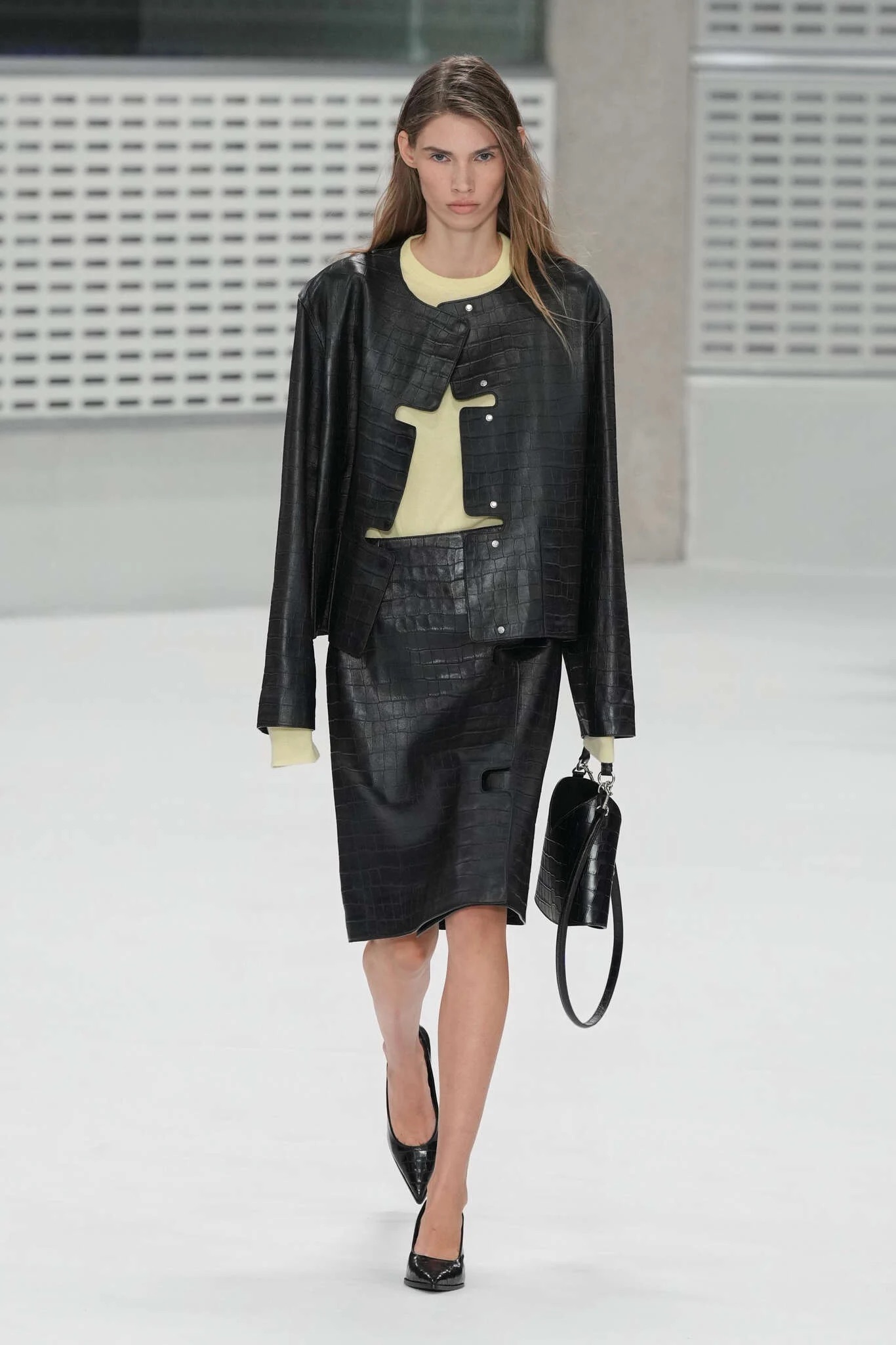 Modelo con un conjunto negro de chaqueta corta y falda midi en piel con textura texturizada, sobre una blusa ligera en tono claro y bolso de mano rígido.