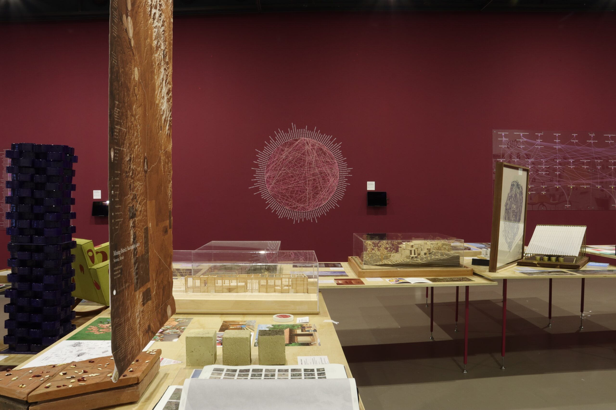 Sala de exposición con paredes en tono burdeos y mesas centrales que exhiben maquetas, muestras de materiales, publicaciones y un gráfico circular tejido con hilos rosas sobre la pared.
