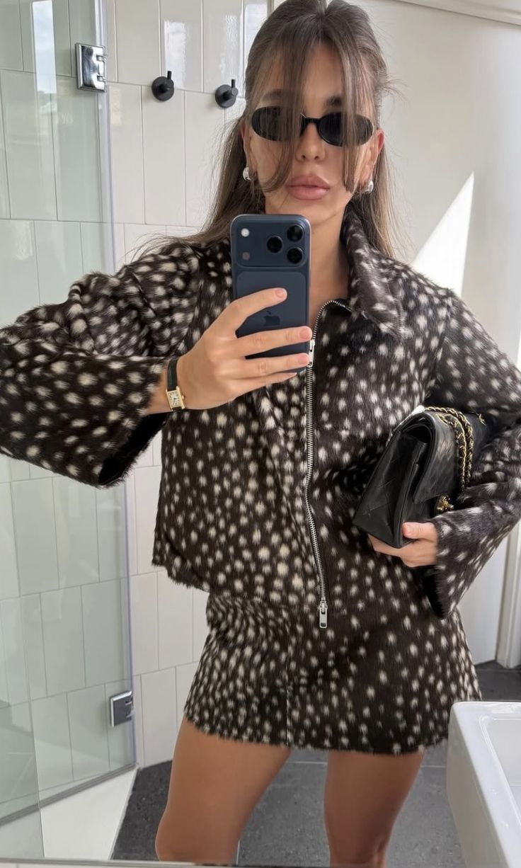 Mujer tomándose una selfie frente al espejo en un baño, luciendo un conjunto de chaqueta con cremallera y minifalda con estampado de cervatillo, gafas de sol oscuras y un bolso acolchado negro.