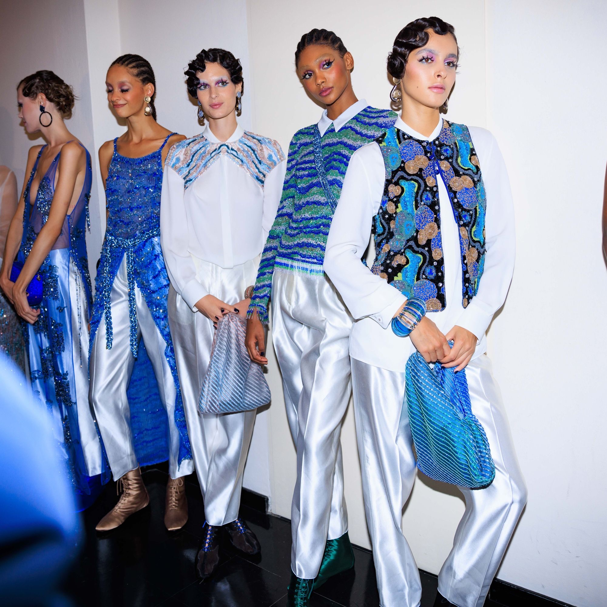 Modelos en el backstage con peinados de ondas al agua vintage y conjuntos en tonos blancos, azules y brillantes con bordados intrincados.