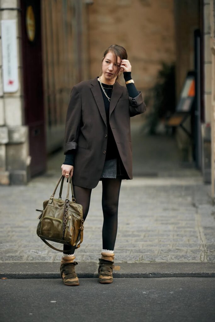 Mujer caminando por la calle vistiendo un blazer oversize marrón oscuro, minifalda, medias negras y zapatillas con plataforma y calcetines cortos visibles.