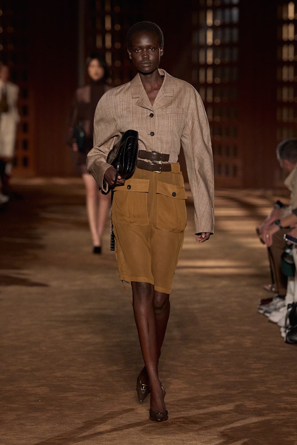 Modelo desfilando con una chaqueta cruzada estructurada en tono marrón claro con hombros marcados, doble cinturón negro en la cintura y una falda midi utility a juego con grandes bolsillos frontales, cortesía de Ferragamo.