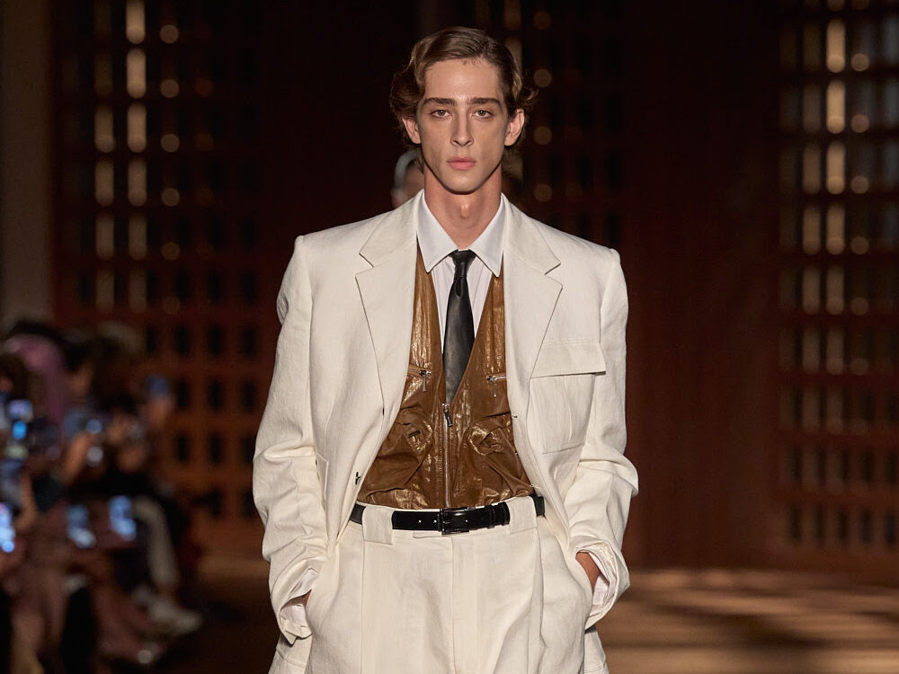 Modelo masculino luciendo un traje sastre en tono blanco marfil de corte relajado, acompañado por una camisa blanca, corbata oscura y un chaleco estilo chaleco táctico de piel en tono marrón con cremalleras, cortesía de Ferragamo.