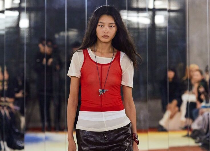 Modelo en la pasarela portando una camiseta corta roja de malla transparente sobre una camiseta blanca de manga corta, combinada con una falda midi de efecto piel.