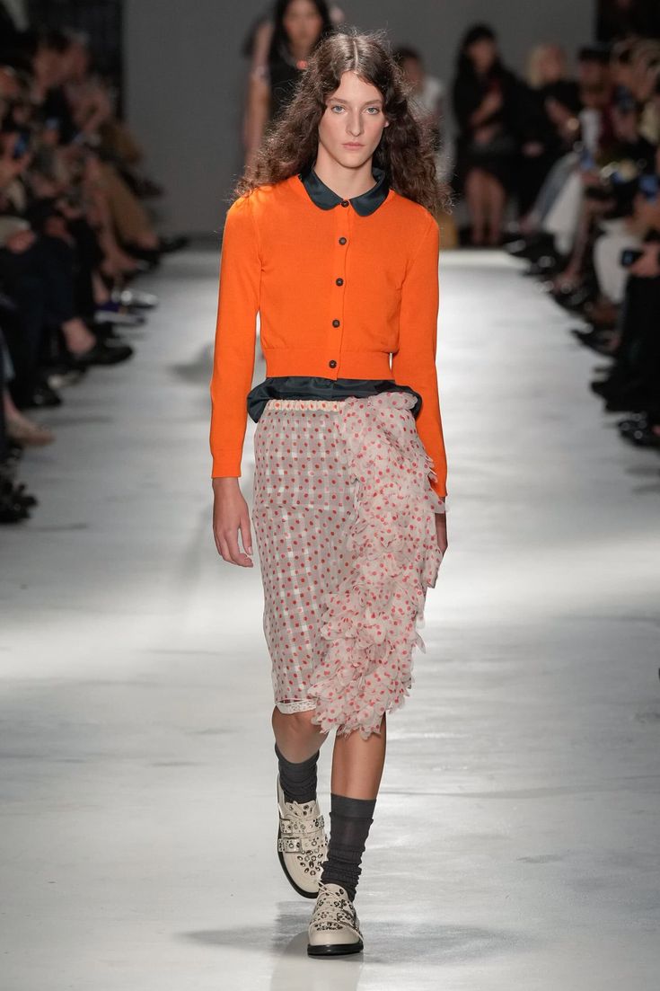 Modelo desfilando con un cárdigan naranja brillante de cuello tipo polo abotonado y una falda midi transparente con estampado de lunares y volantes, acompañada de calcetines oscuros y zapatos claros.